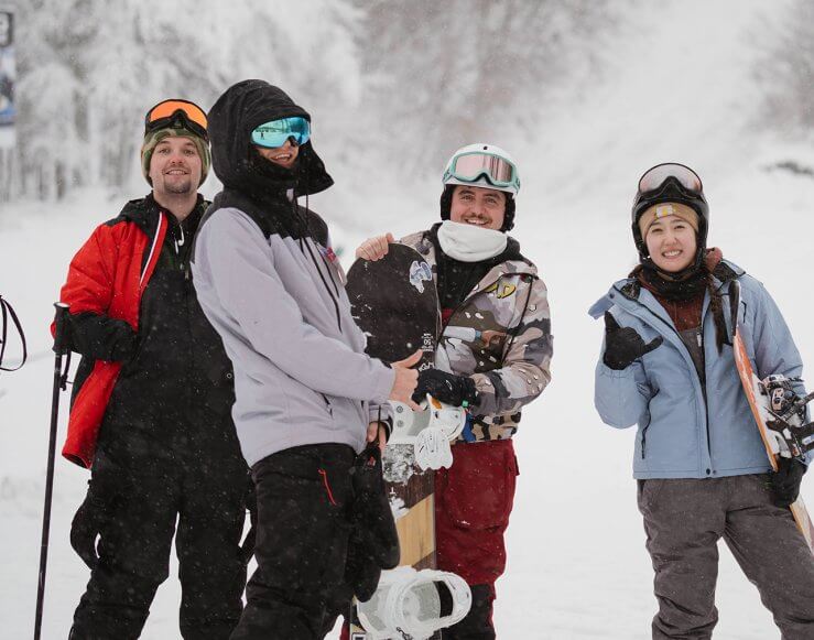 4 Snowboarders smiling for the camera next to the yellow chairlift with a snowy background